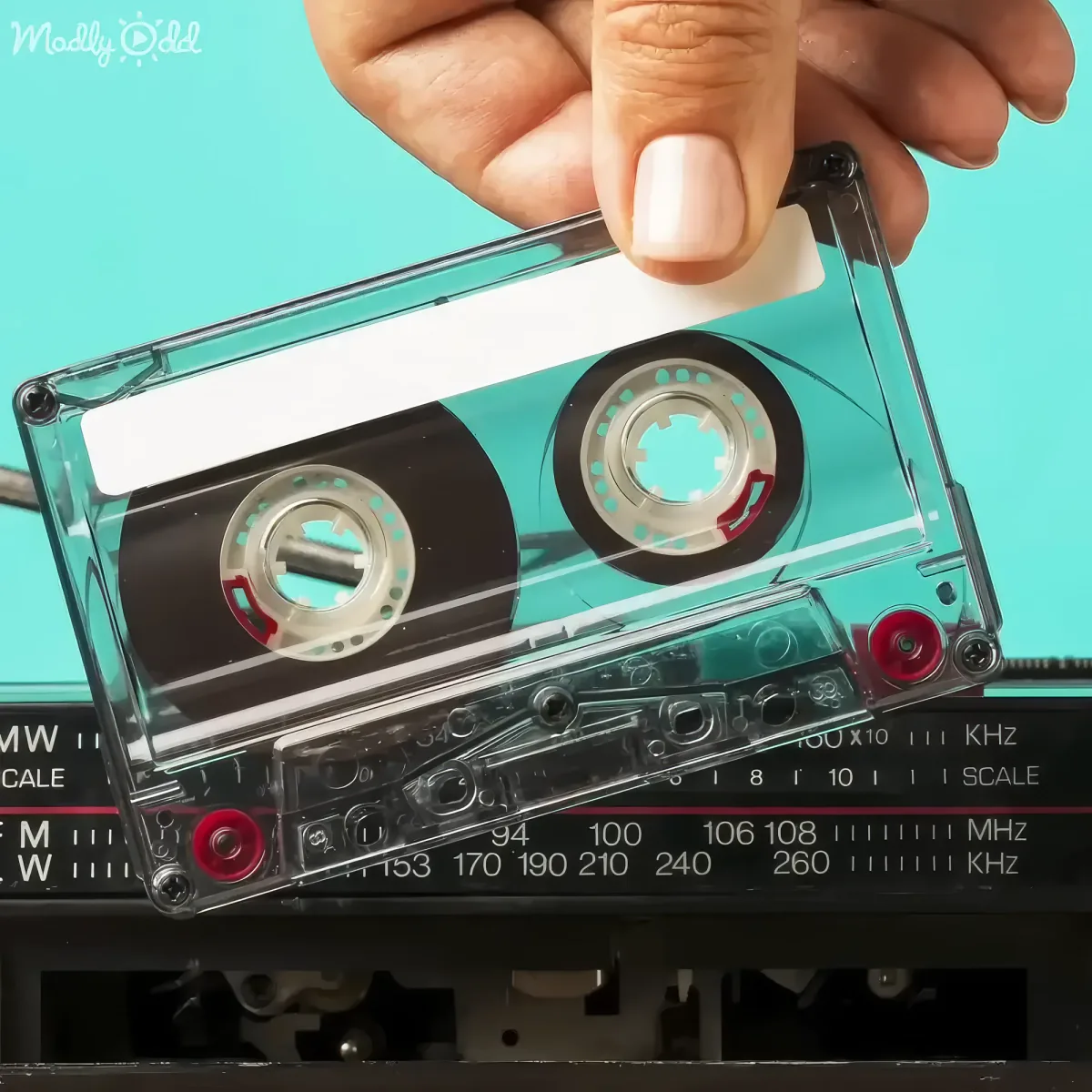A hand holds a transparent cassette tape over a radio dial with MHz and kHz scales.