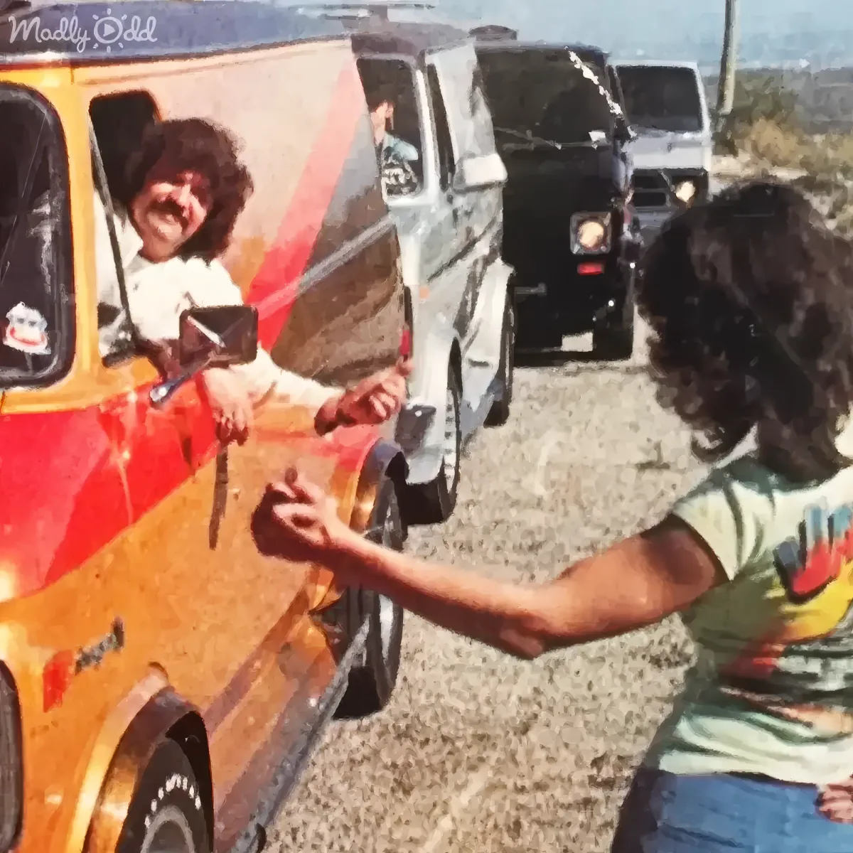 A man with curly hair shakes hands with another man in front of a colorful van on a gravel road.