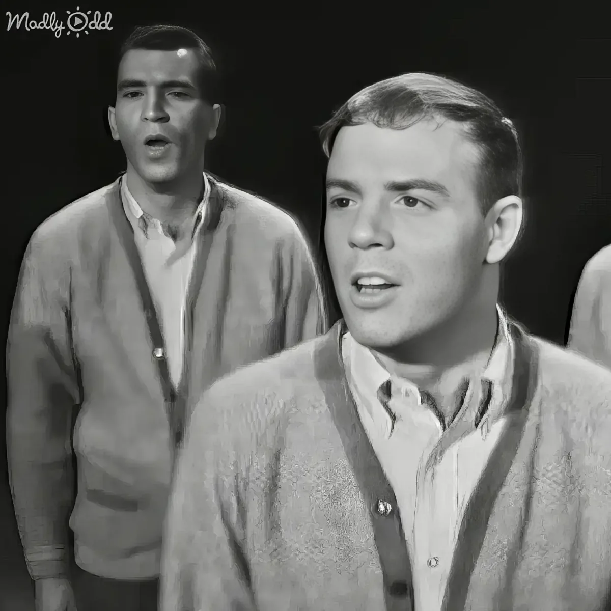 A man in a textured sweater sings, with another man in a cardigan behind him, both in a black-and-white setting.