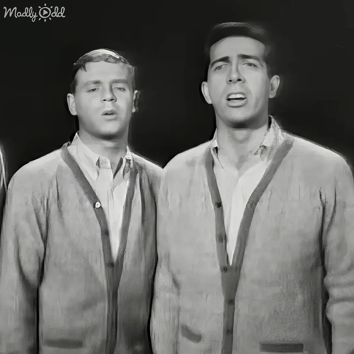 Two men in matching cardigans and collared shirts stand side by side, possibly singing or speaking.