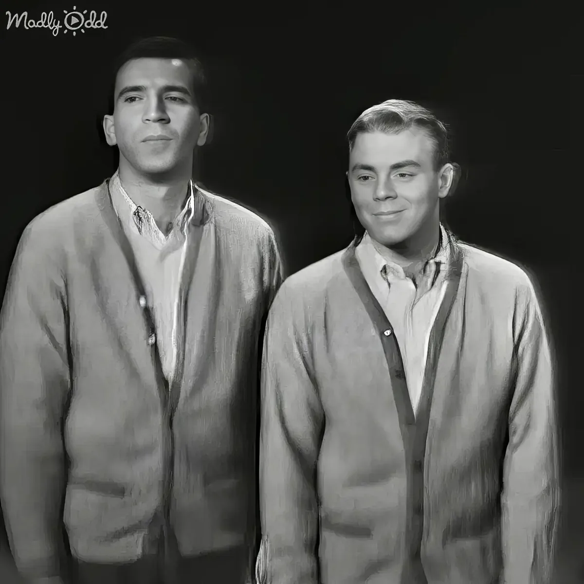Two men in vintage attire stand side by side, one smiling, both wearing open cardigans over collared shirts.
