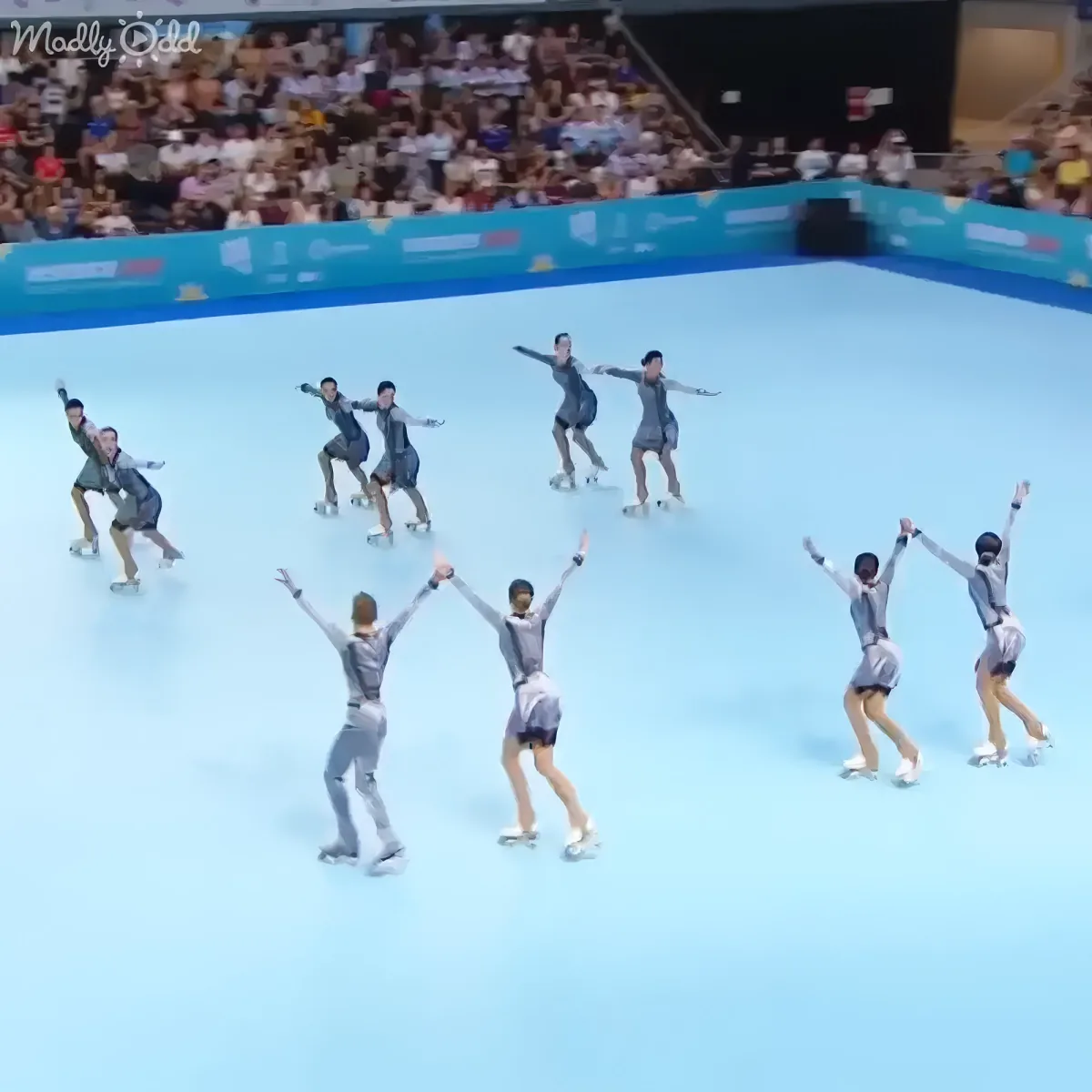 Skaters in synchronized formation on ice, arms raised, performing in front of a crowd.