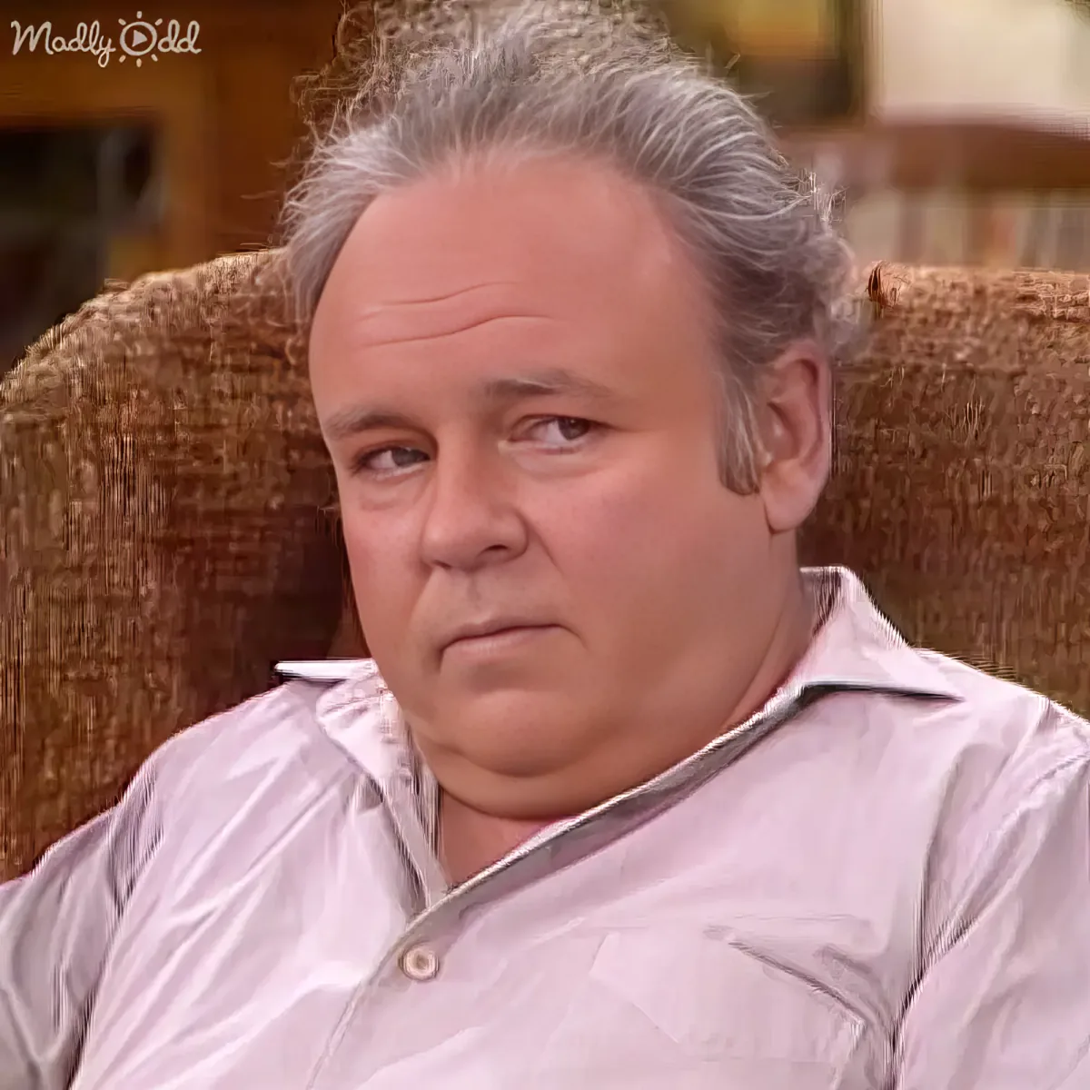 A man with gray hair sits in a brown chair, wearing a light-colored shirt, looking slightly to the side.