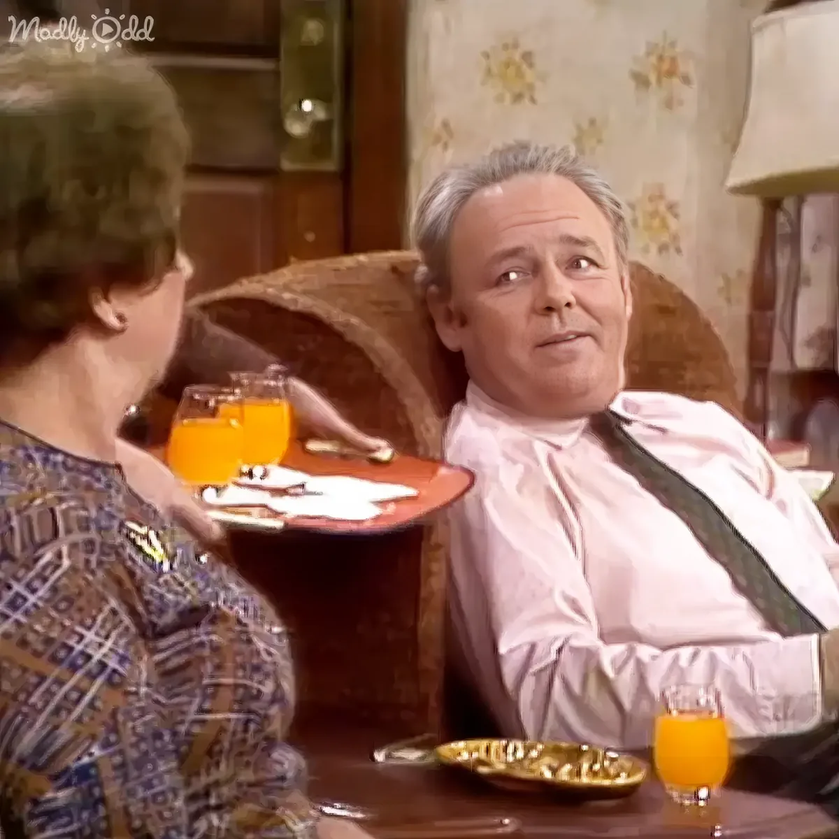 A man in a pink shirt and suspenders sits in a chair, holding a tray with orange juice and cookies, while a woman in a...
