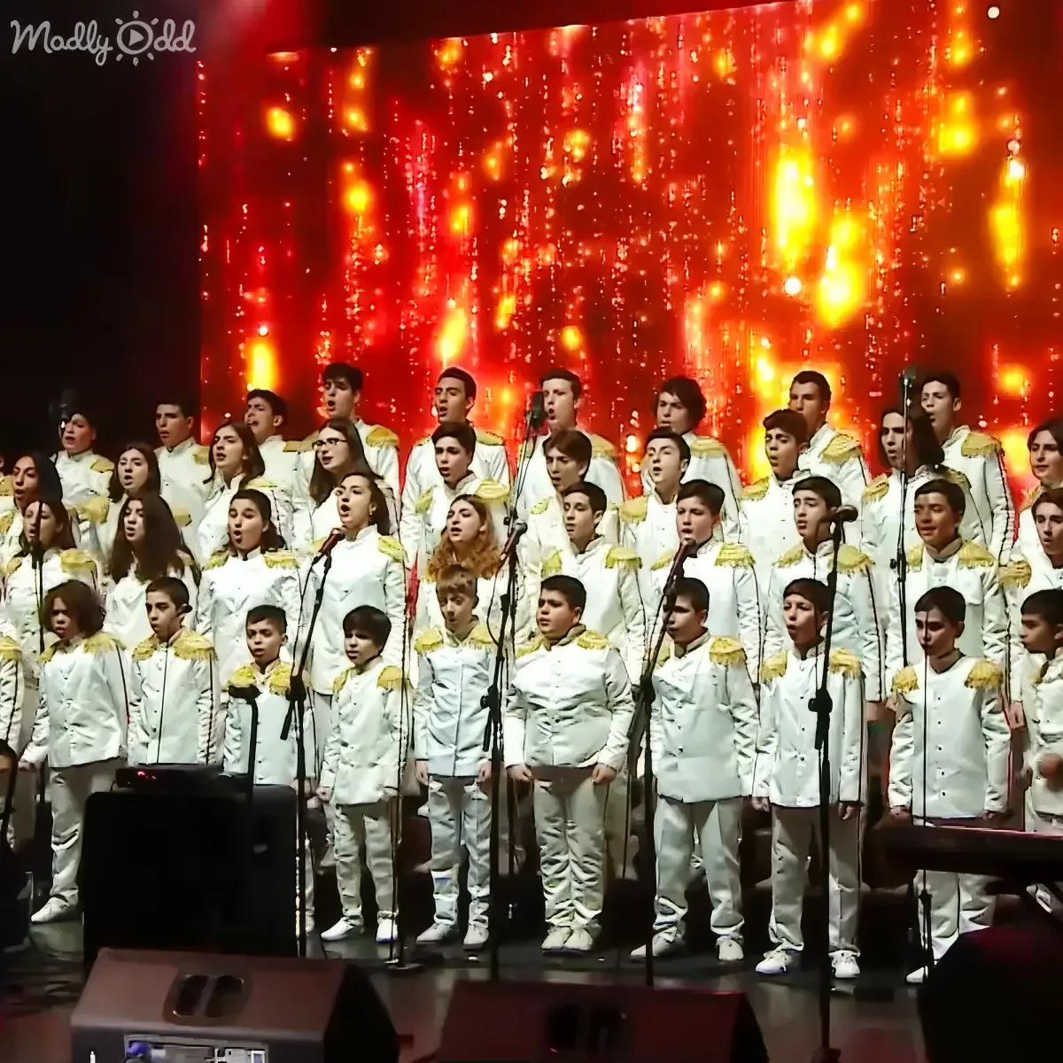A choir in white uniforms sings on stage with a vibrant red backdrop and falling lights.