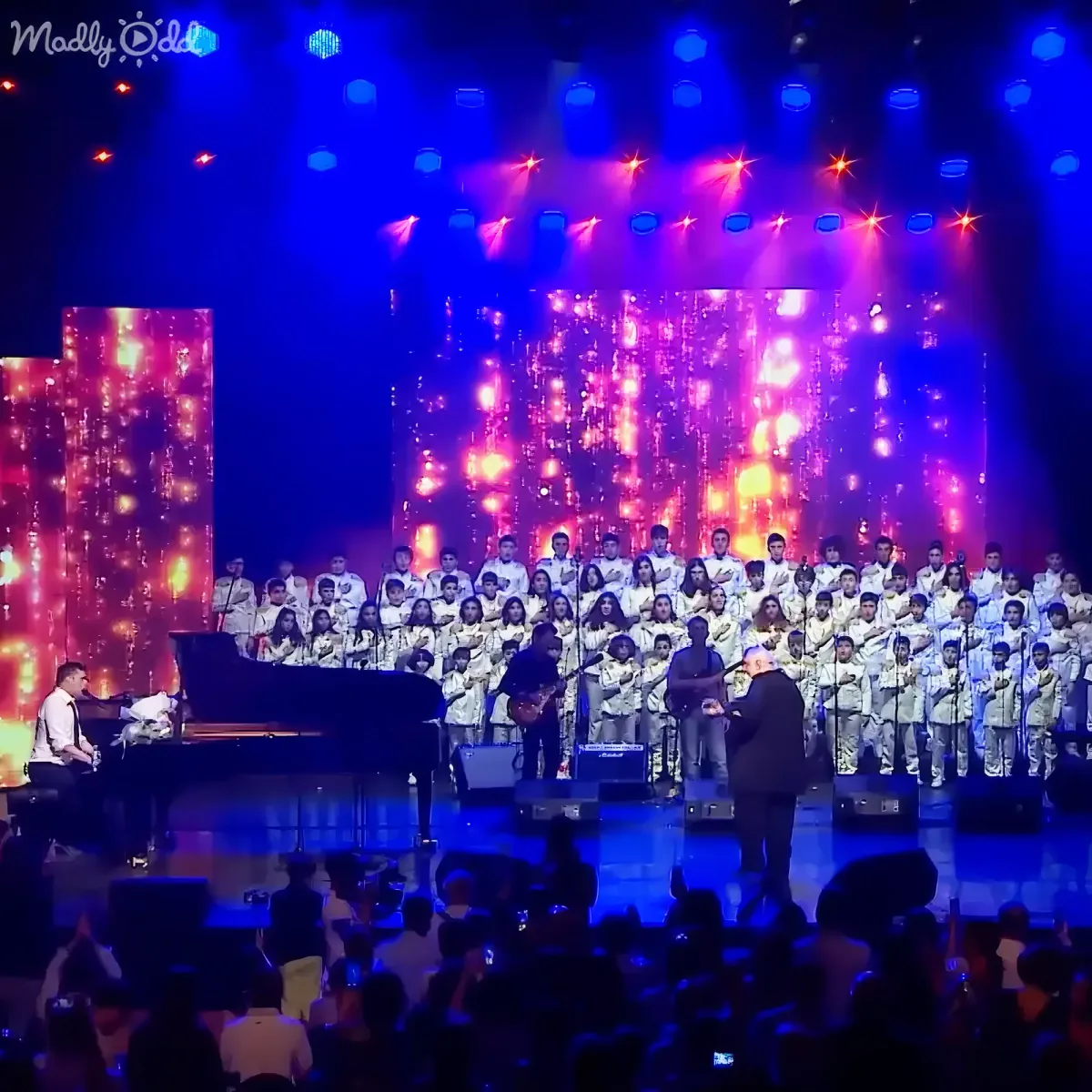 A grand piano, guitar, and conductor lead a large choir on a stage with vibrant lights.