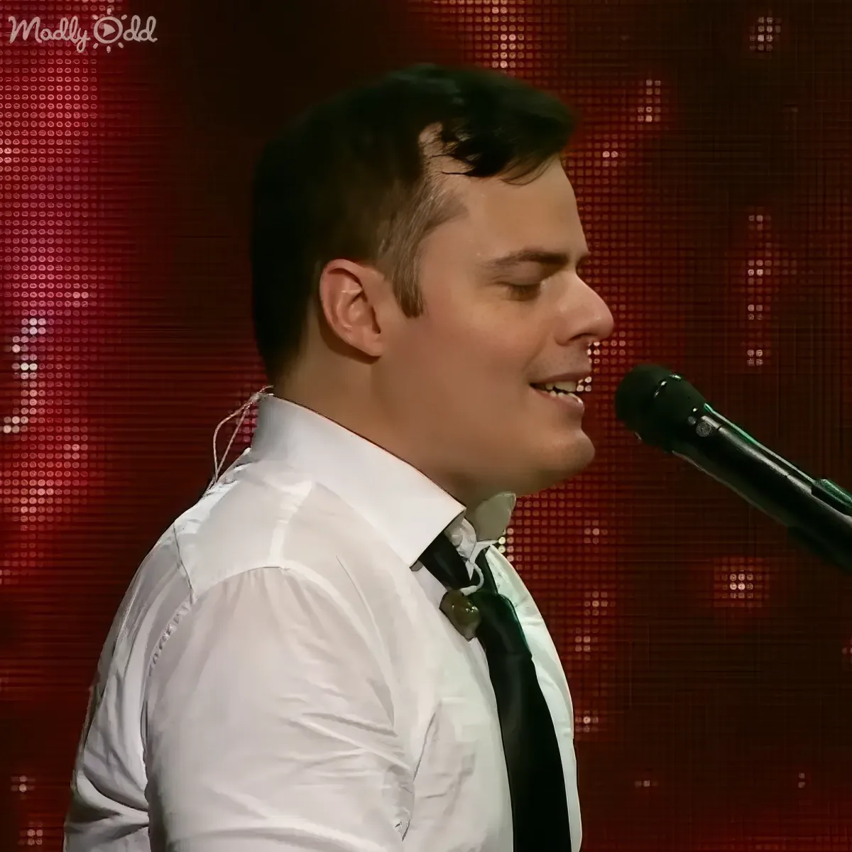 A man in a white shirt and black tie sings into a microphone against a red, pixelated background.