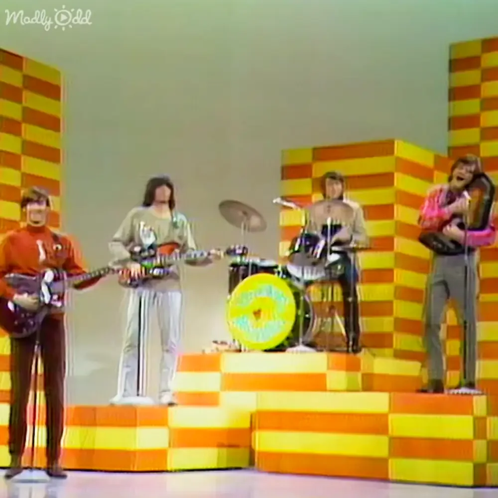 A band performs on a stage with vibrant orange and yellow striped blocks in the background.