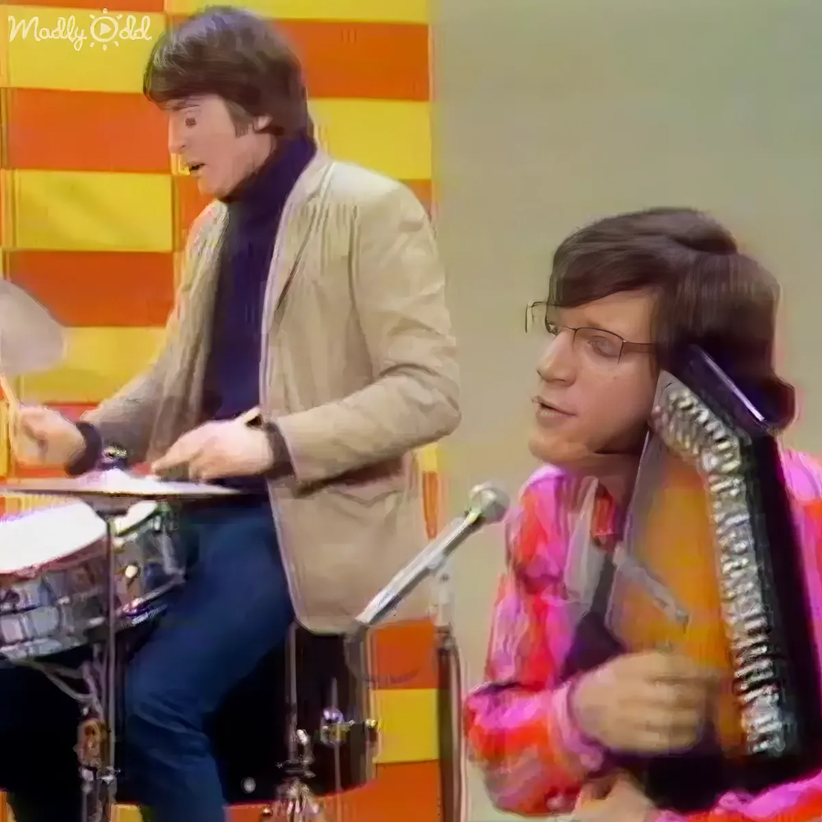 A man in a beige jacket plays drums while another in a pink shirt holds a guitar.