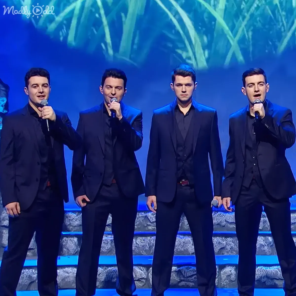 Four men in black suits sing on stage with microphones, illuminated by blue lights and a green leafy backdrop.