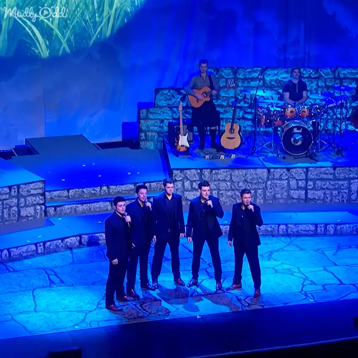A group of five men in black suits sing on stage, with musicians playing instruments behind them.
