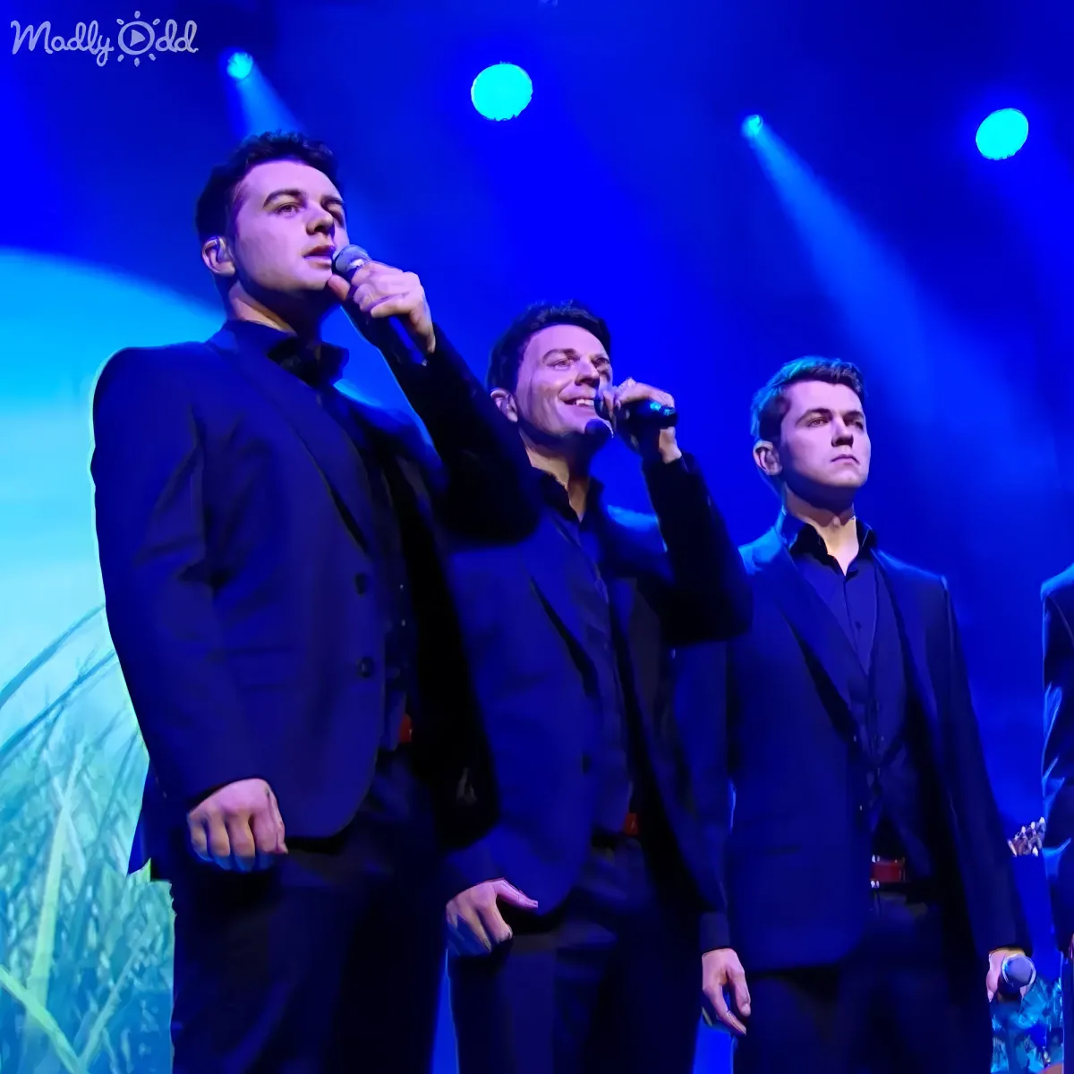 Three men in black suits sing on stage under blue lights, one holding a microphone.