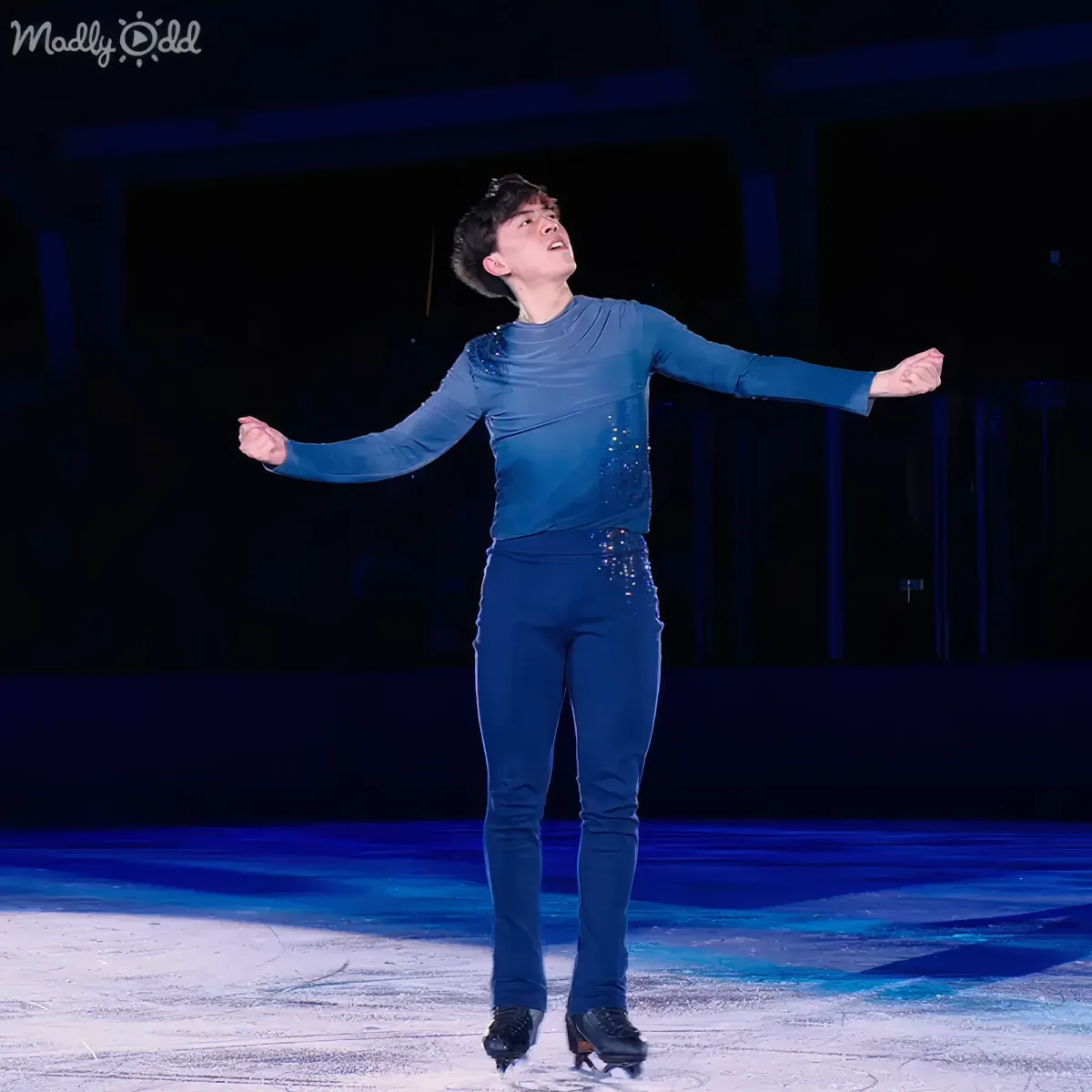 A figure skater in a blue outfit stands gracefully on ice with arms outstretched.