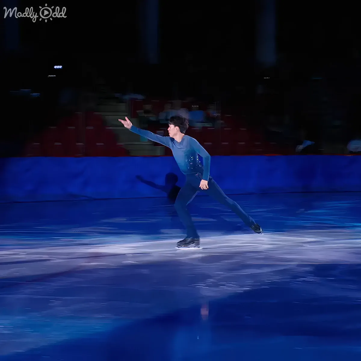 A figure skater in a blue outfit glides gracefully on ice, arms extended, with a blurred audience in the background.