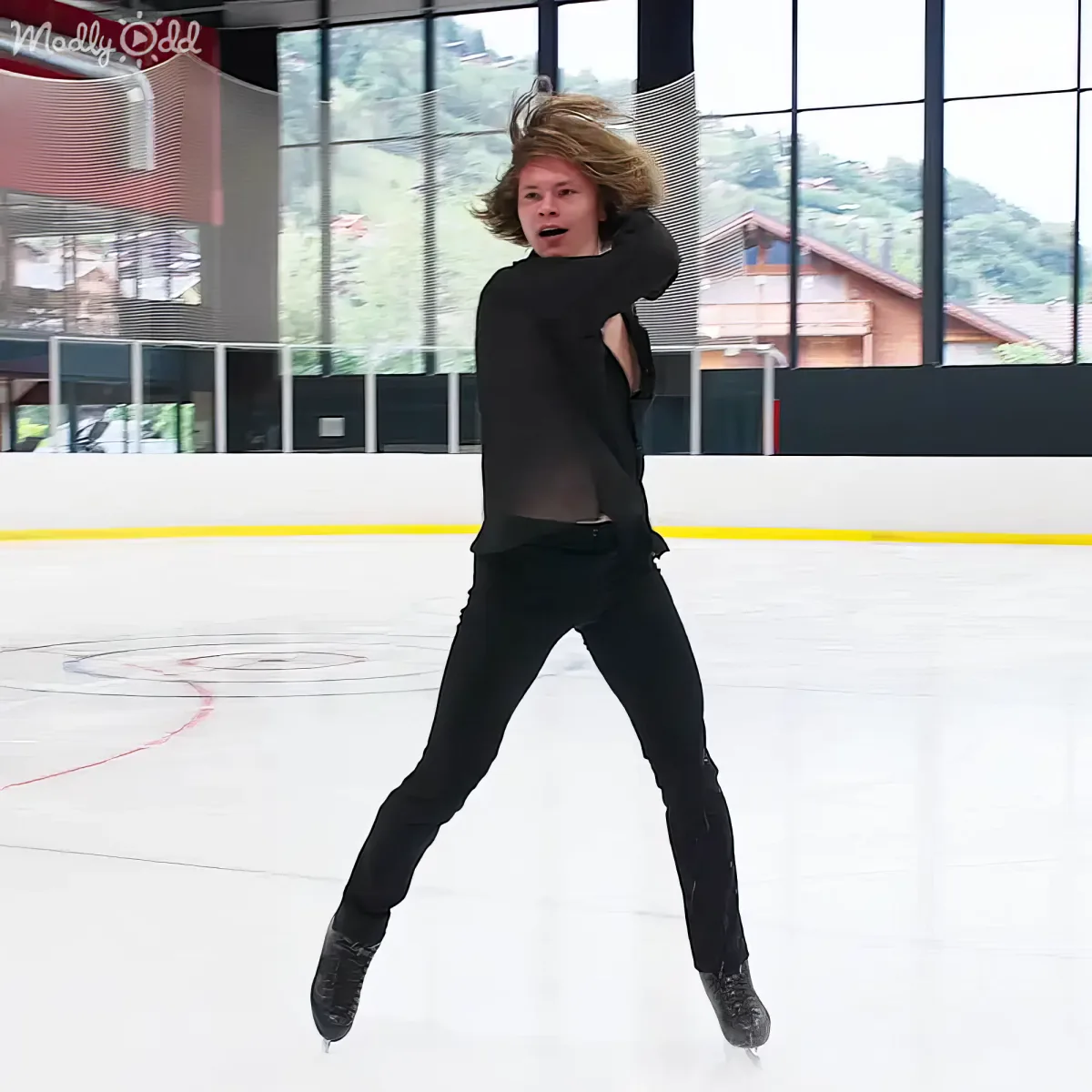 A person in black attire skates on an ice rink, with a scenic background of buildings and trees.