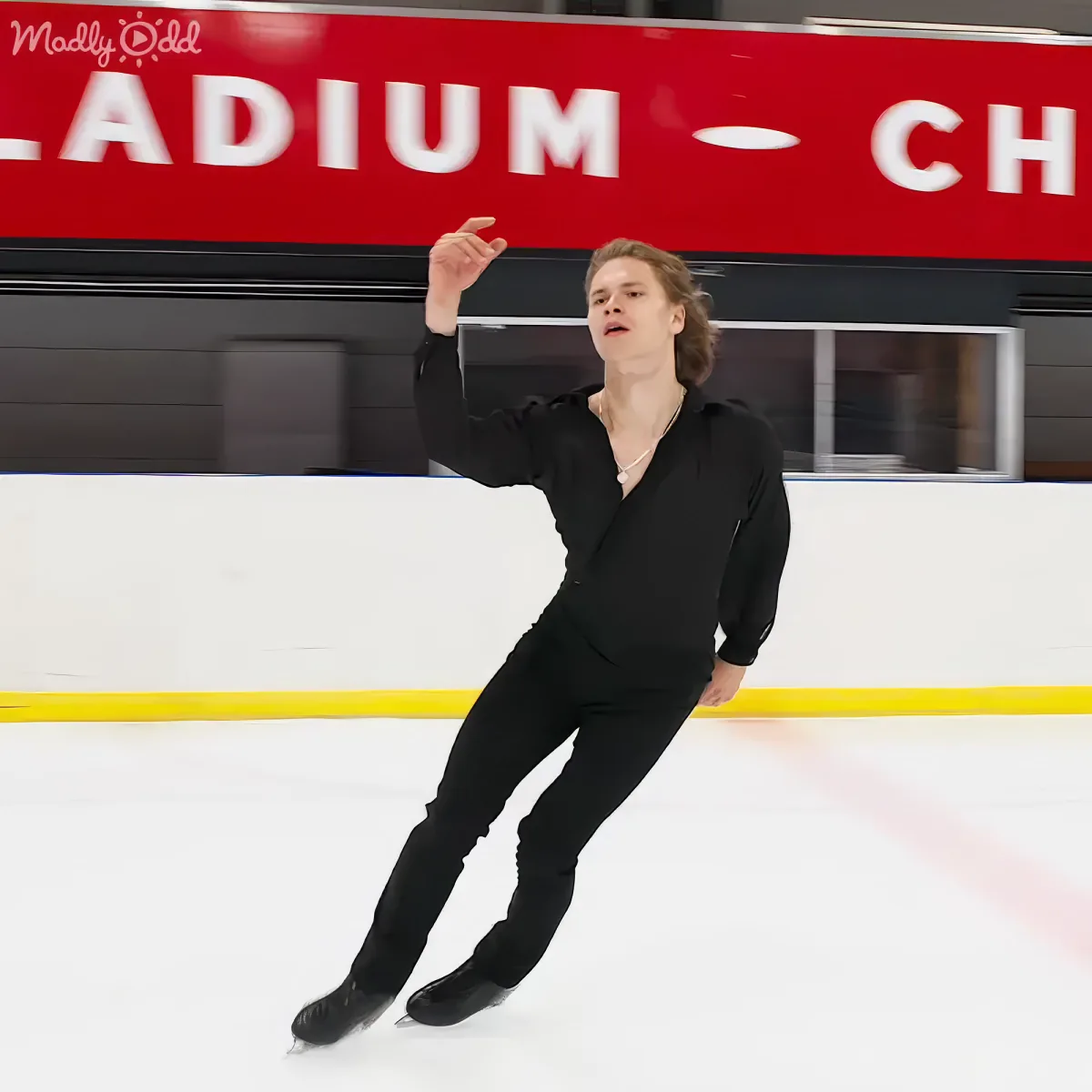 A person in black attire skates on ice, arms outstretched, with a red sign in the background.