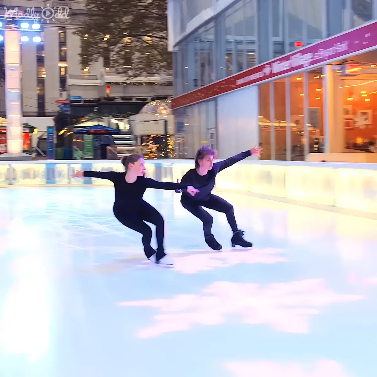 Two figure skaters in black perform a synchronized move on an ice rink with a festive background.