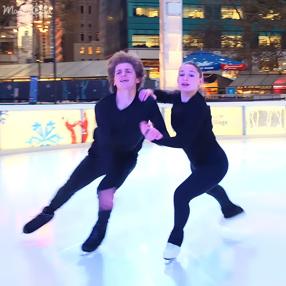 Two people in black outfits skate together on an ice rink, holding hands and smiling.