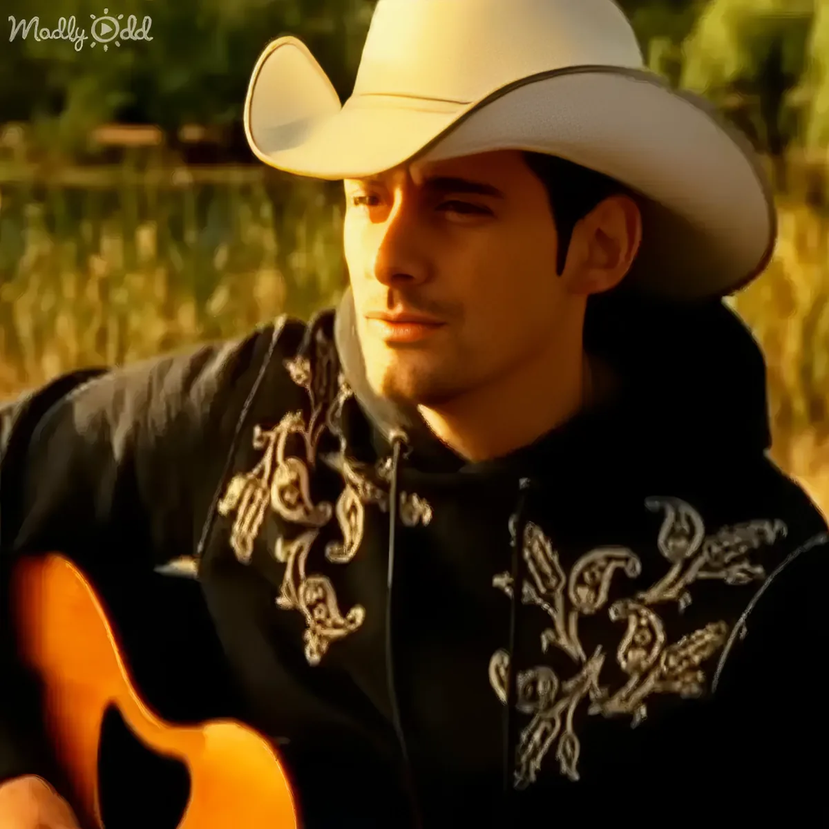 A man in a cowboy hat and embroidered shirt holds a guitar, set against a blurred natural background.
