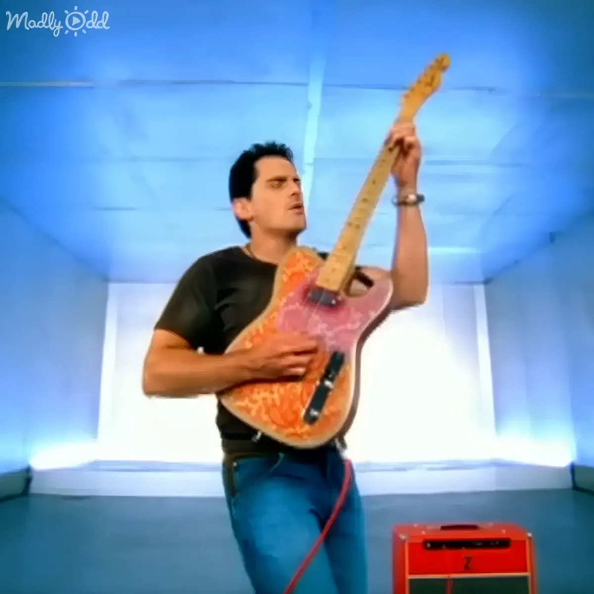 A man in a black shirt and blue jeans plays a pink and orange guitar on stage.