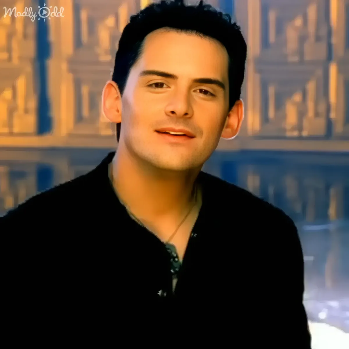 A man in a black shirt with a necklace, smiling slightly, against a blurred background of a building.