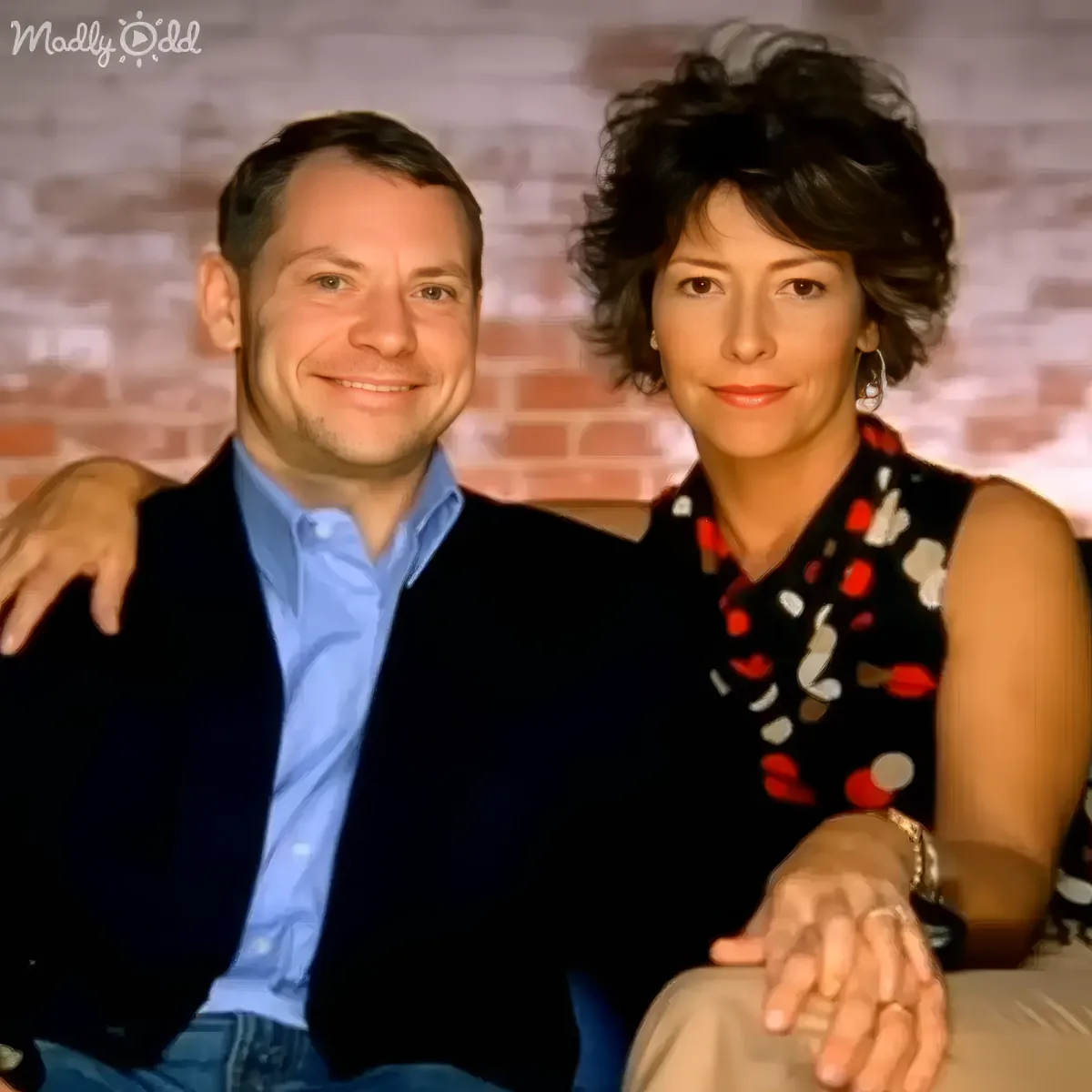 A man and woman are sitting together, smiling, with a brick wall background.