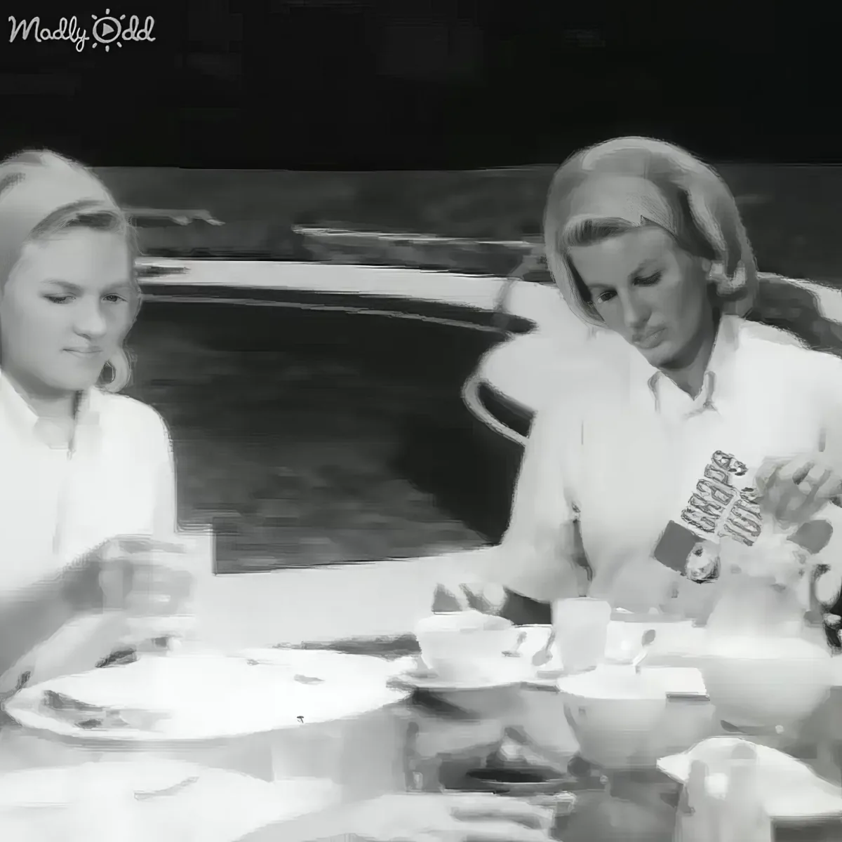 Two individuals are seated at a table with tea cups, engaged in conversation, with a car visible in the background.