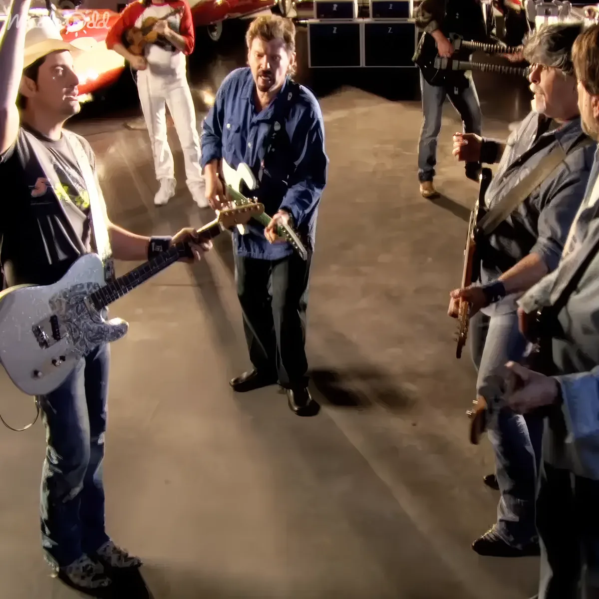 A man in a blue shirt and jeans plays guitar, surrounded by other musicians in a garage setting.