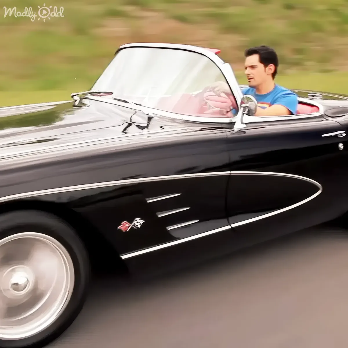 A man drives a classic black Corvette convertible on a road, wearing a blue shirt.