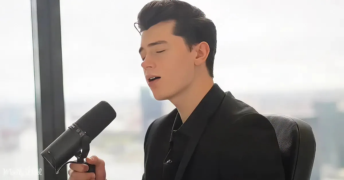 A person in a black suit holds a microphone, seated in front of a window with a cityscape view.