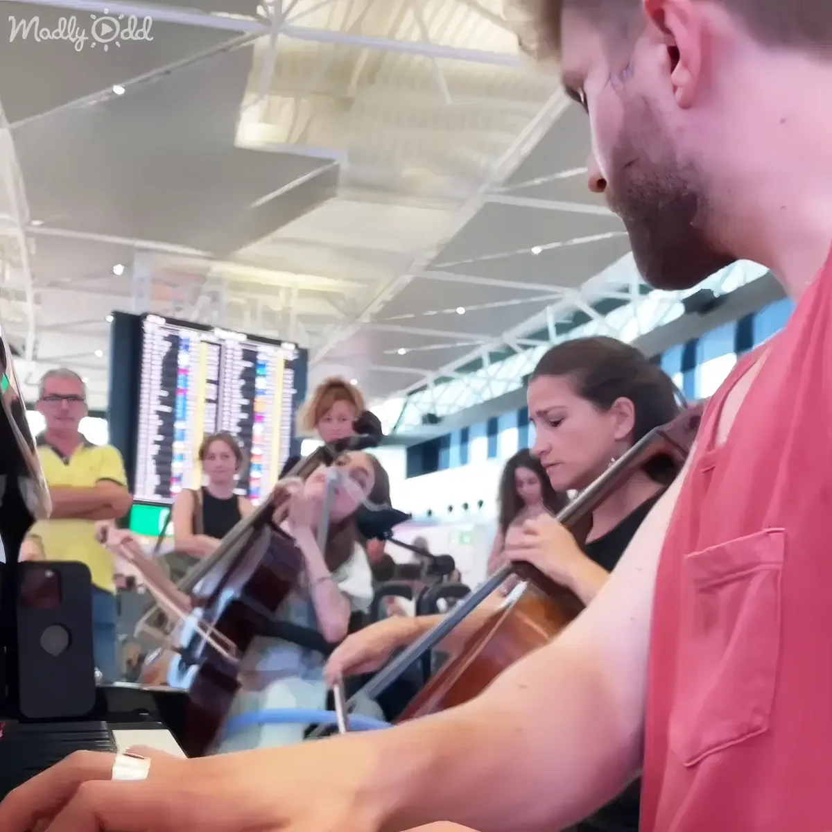 A man in a pink shirt plays the cello in a busy airport, surrounded by other musicians and travelers.