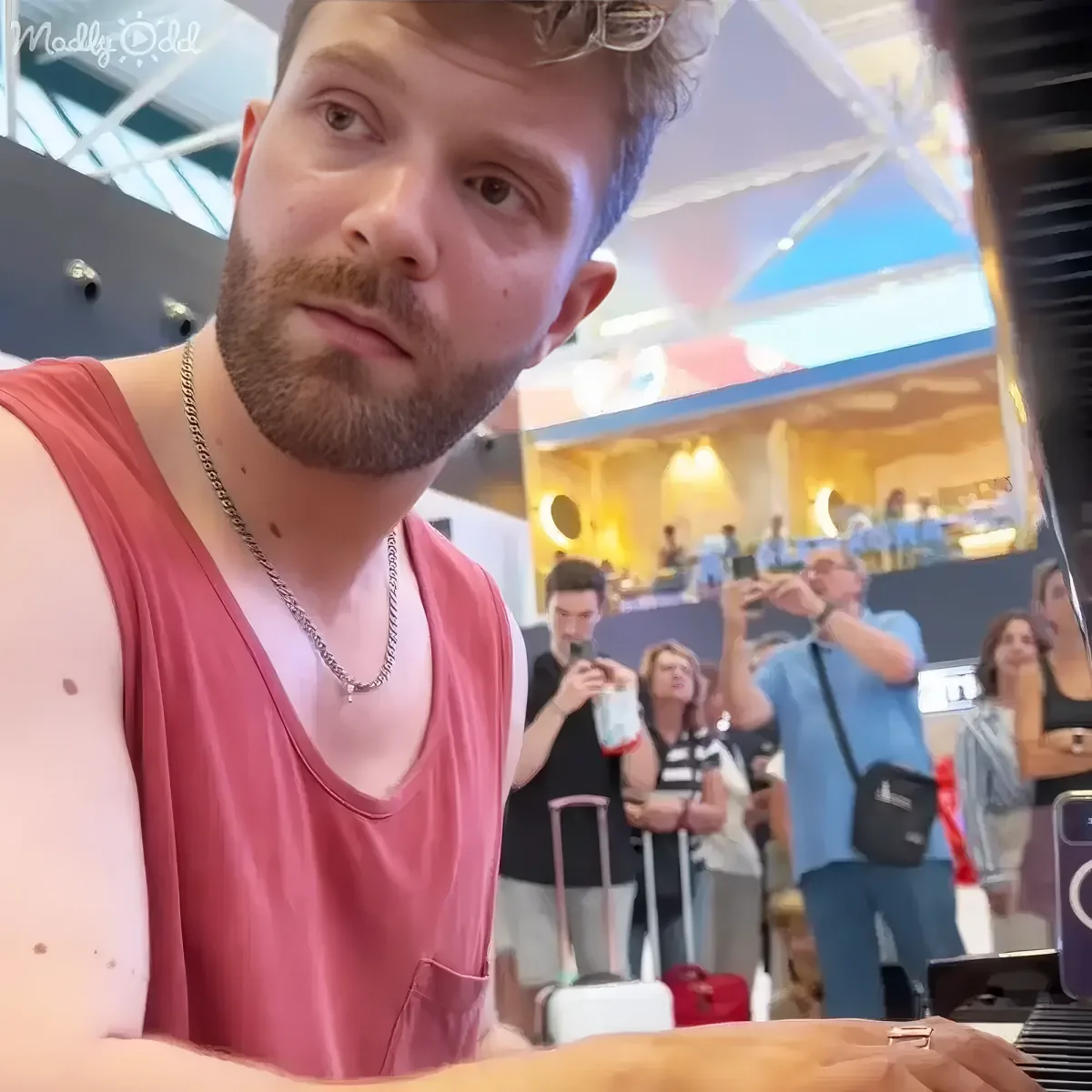 A man in a red tank top and necklace is taking a selfie in a busy indoor setting with people around him.