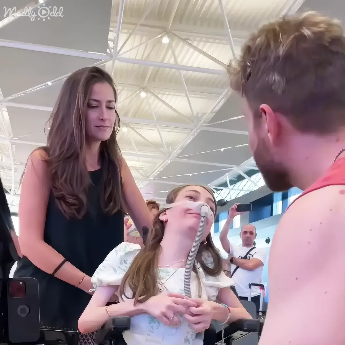 A woman in a wheelchair, wearing a floral shirt and an oxygen mask, is surrounded by people in a bright, open space.