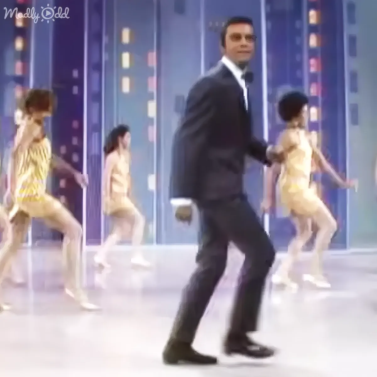 A man in a suit dances with children in gold outfits on a stage with colorful lights.