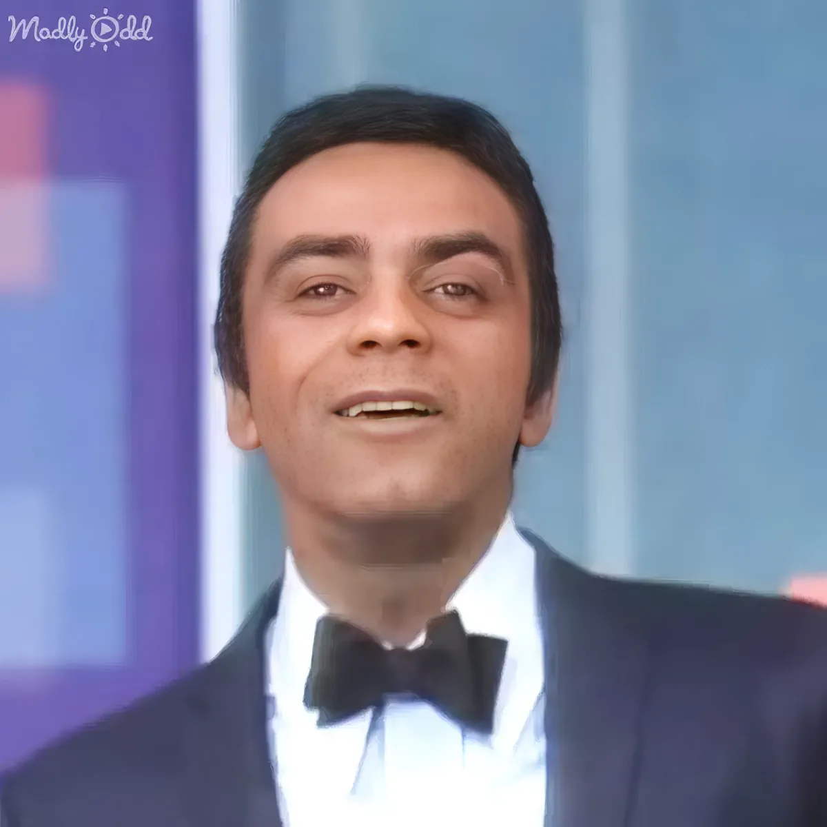 A man in a tuxedo with a bow tie is smiling, wearing a white shirt, against a blurred background.