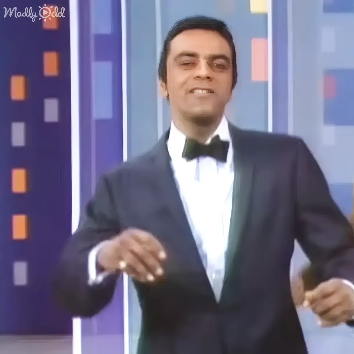 A man in a tuxedo stands confidently with hands slightly raised, wearing a bow tie and a ring, against a colorful backdrop.