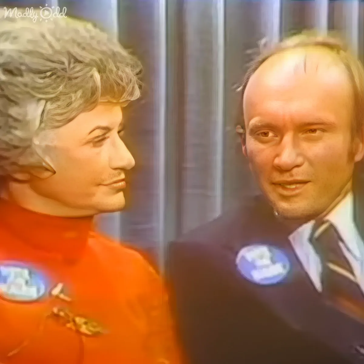 A man in a suit with a badge and a woman in an orange outfit are standing close, seemingly engaged in conversation.