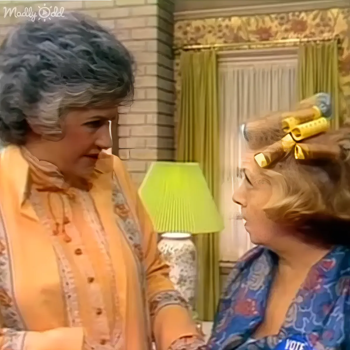 An older woman with short gray hair stands beside a younger woman with blonde hair in curlers, both in a room with a...