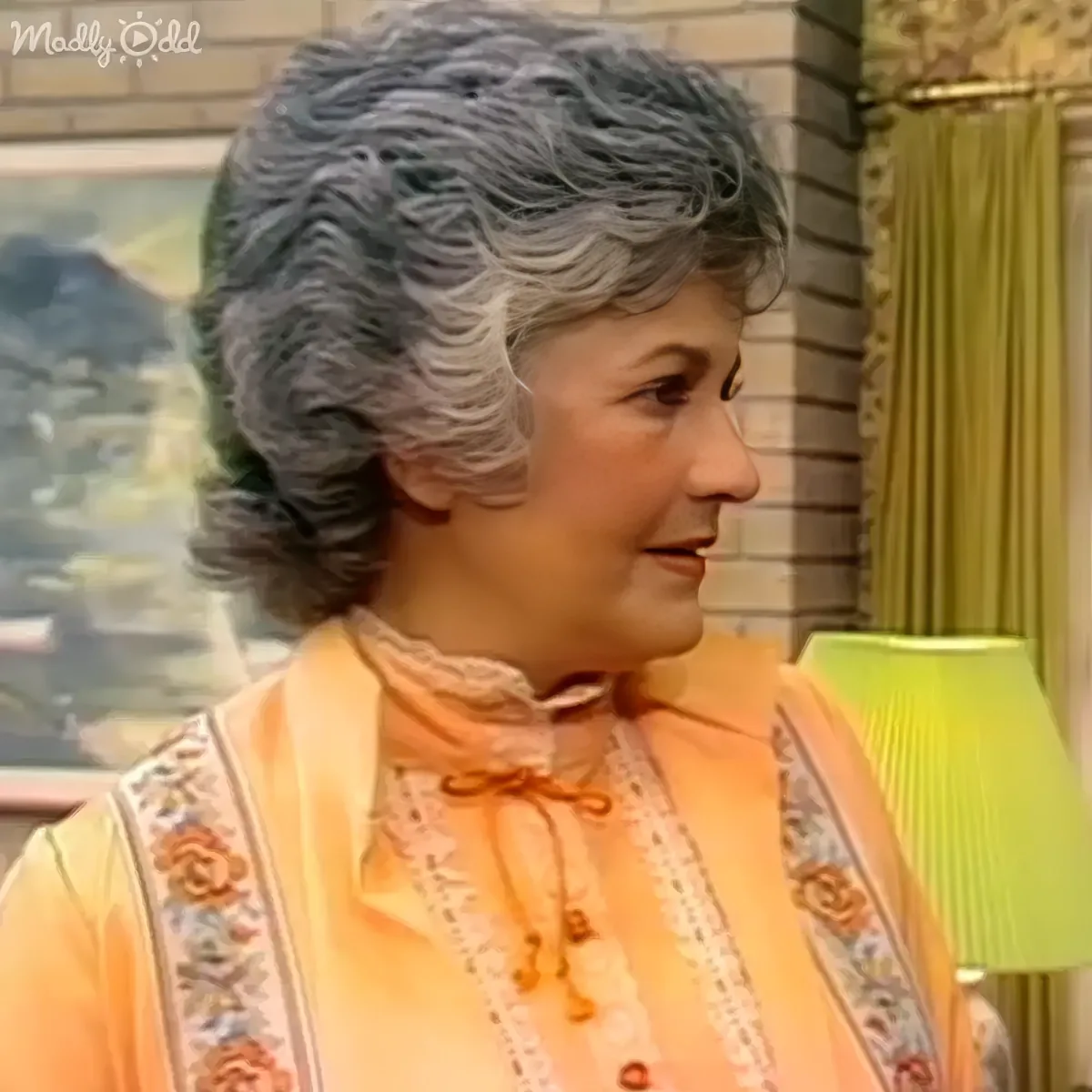 A woman with gray hair in a vintage-style orange dress with lace and floral patterns is smiling.