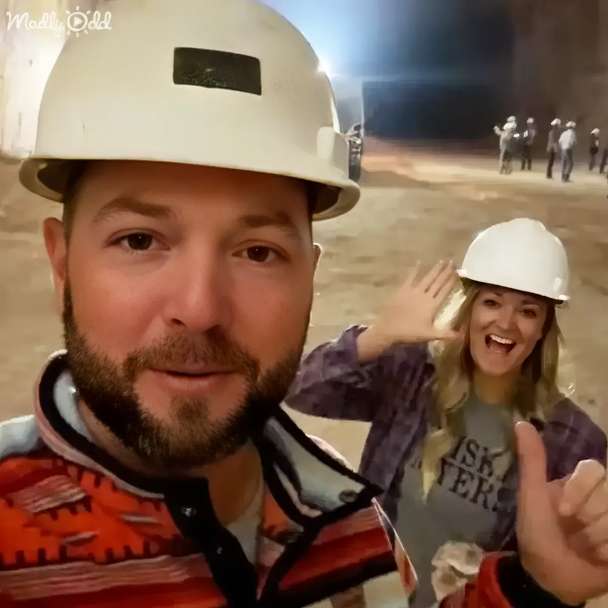 A man in a hard hat and safety vest takes a selfie with a woman in the background, both smiling and waving.