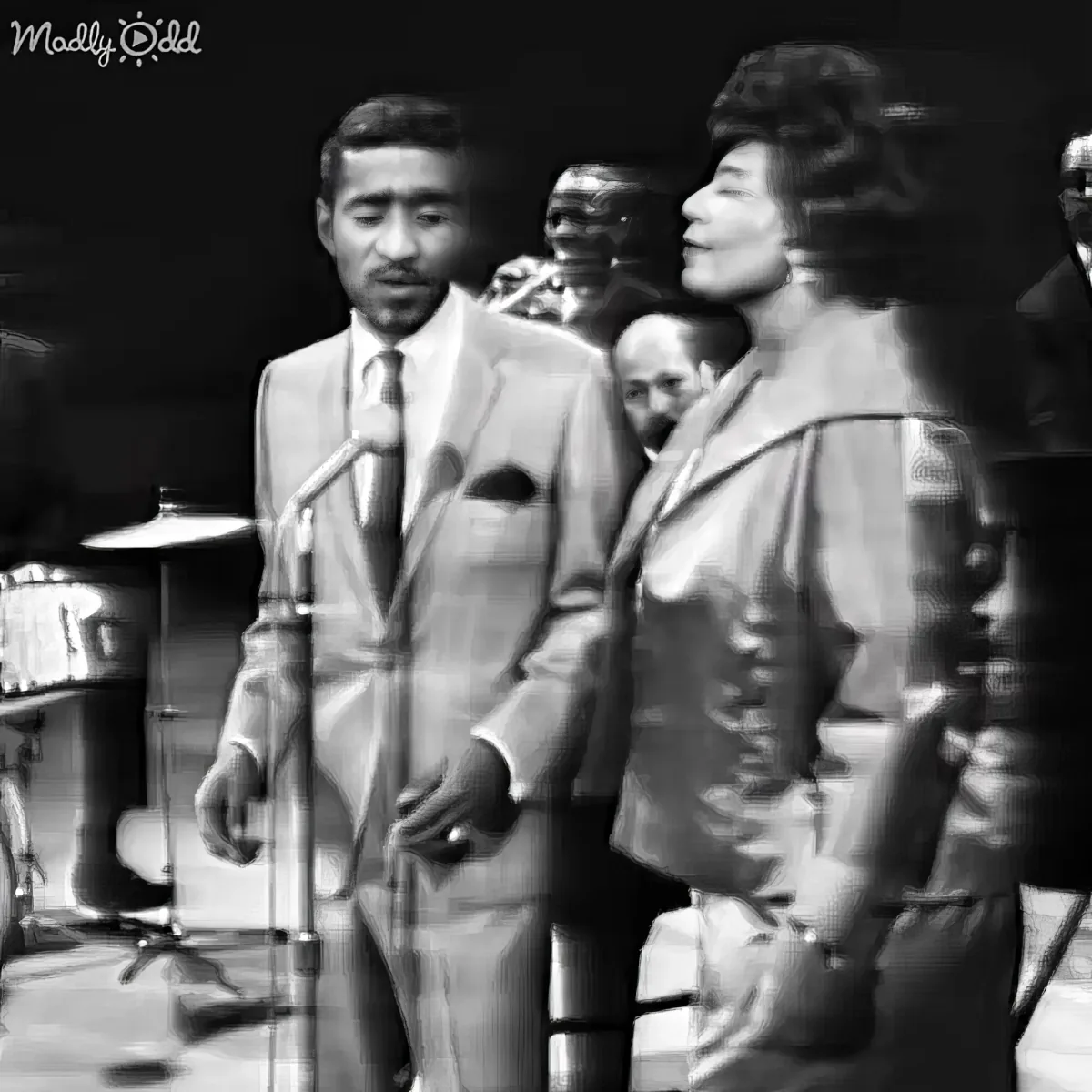 A man in a suit sings into a microphone, accompanied by a woman in a dress, with a band in the background.
