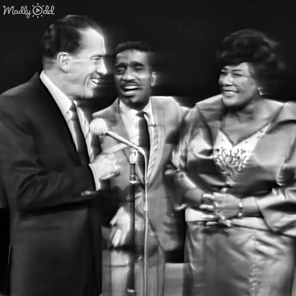 A man in a suit and tie is holding a microphone, smiling, with two other people, one in a suit and the other in a dress...