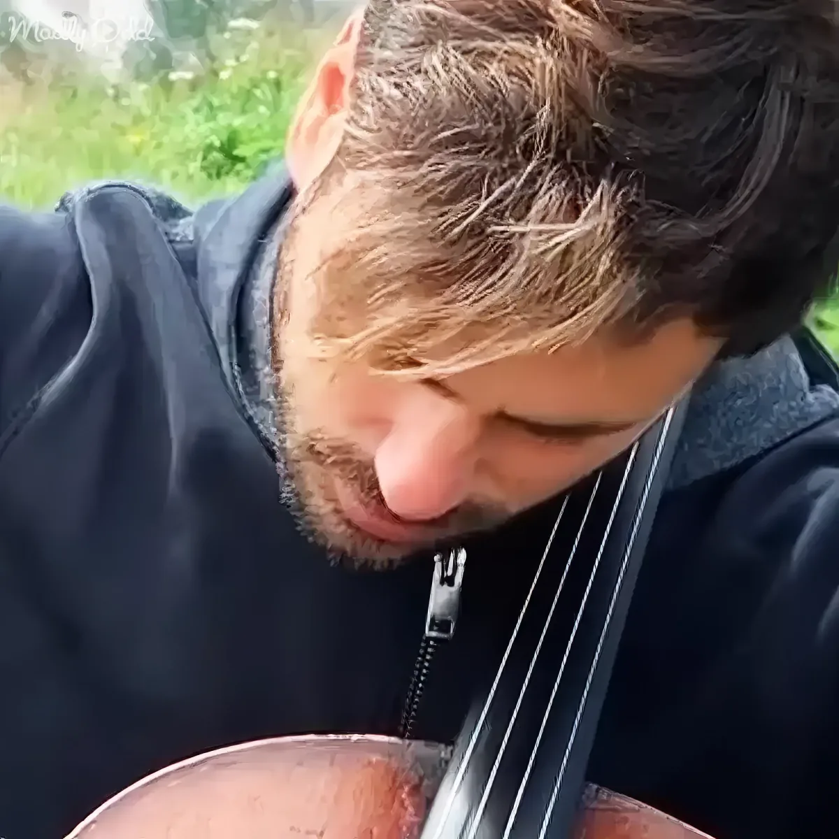 A man with short hair and a beard is playing a cello outdoors, wearing a dark jacket.