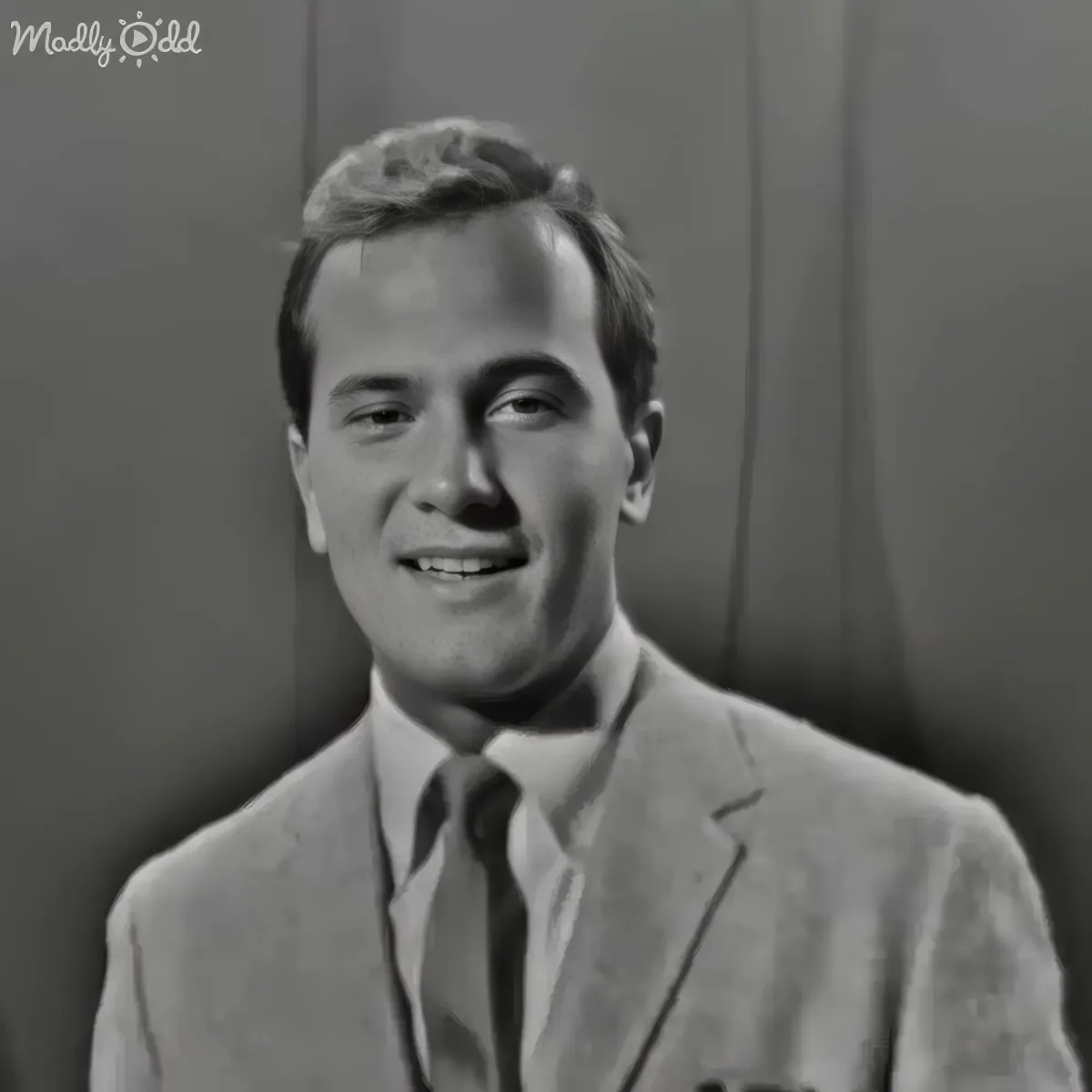 A man in a suit and tie smiles, wearing a light-colored suit jacket and tie, with a neutral background.