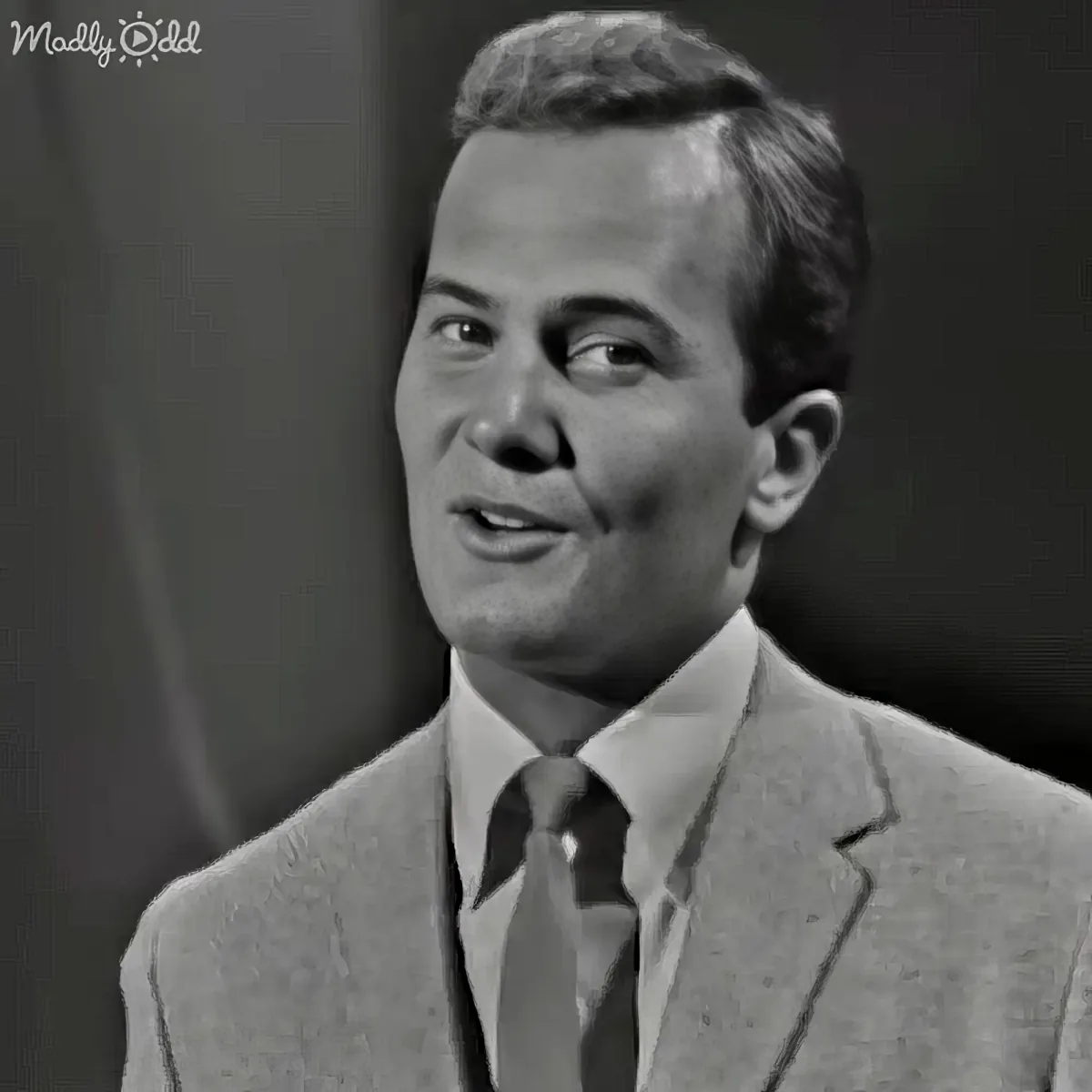 A man in a suit and tie, smiling, with short hair, against a dark background.