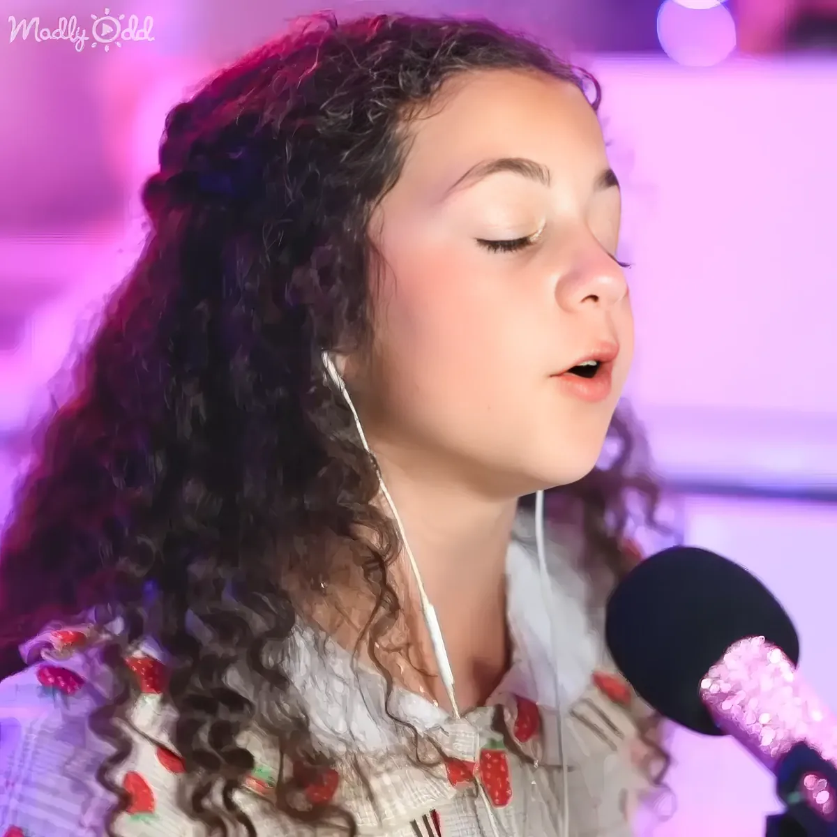 A young girl with long, curly hair sings into a microphone, wearing a white shirt with strawberry patterns.