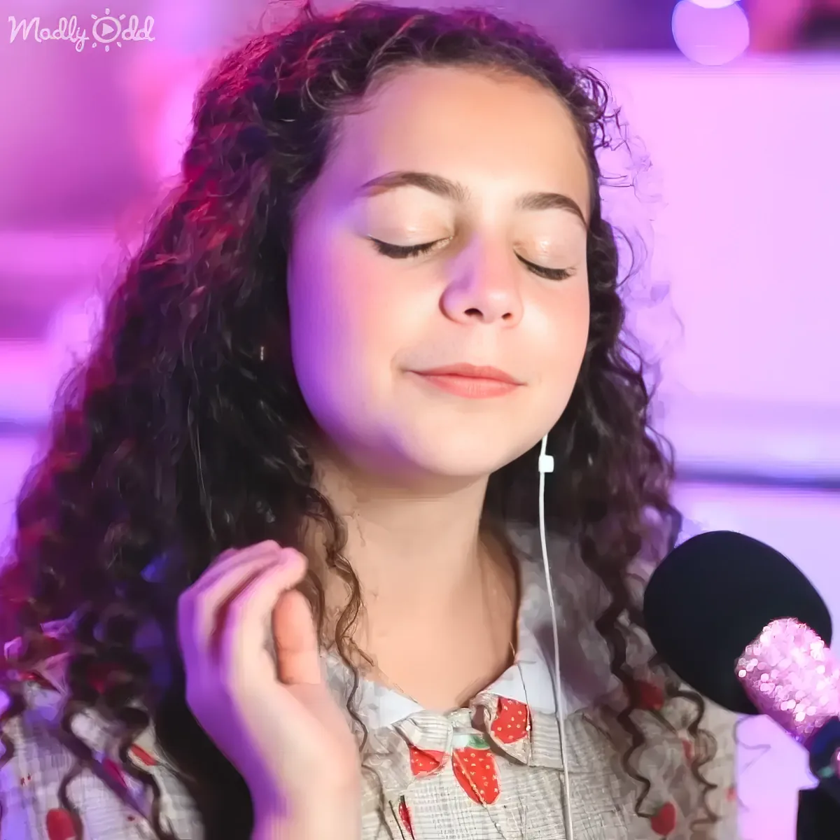 A young person with curly hair, wearing a plaid shirt with strawberry patterns, is holding a microphone and appears to be...