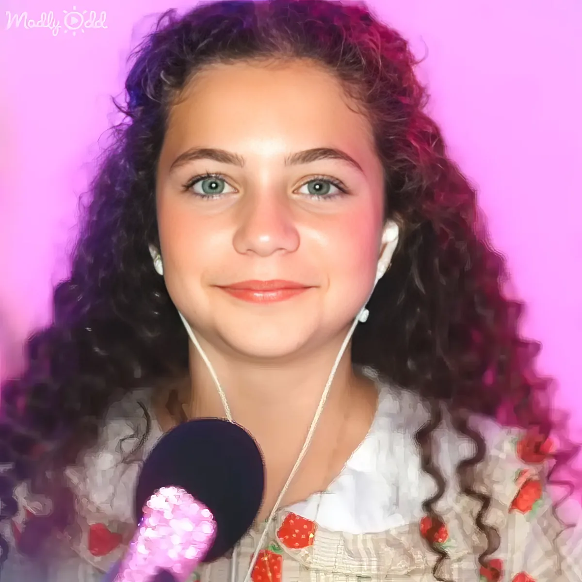 A young person with curly hair and earphones, wearing a strawberry-patterned top, smiles against a pink background.