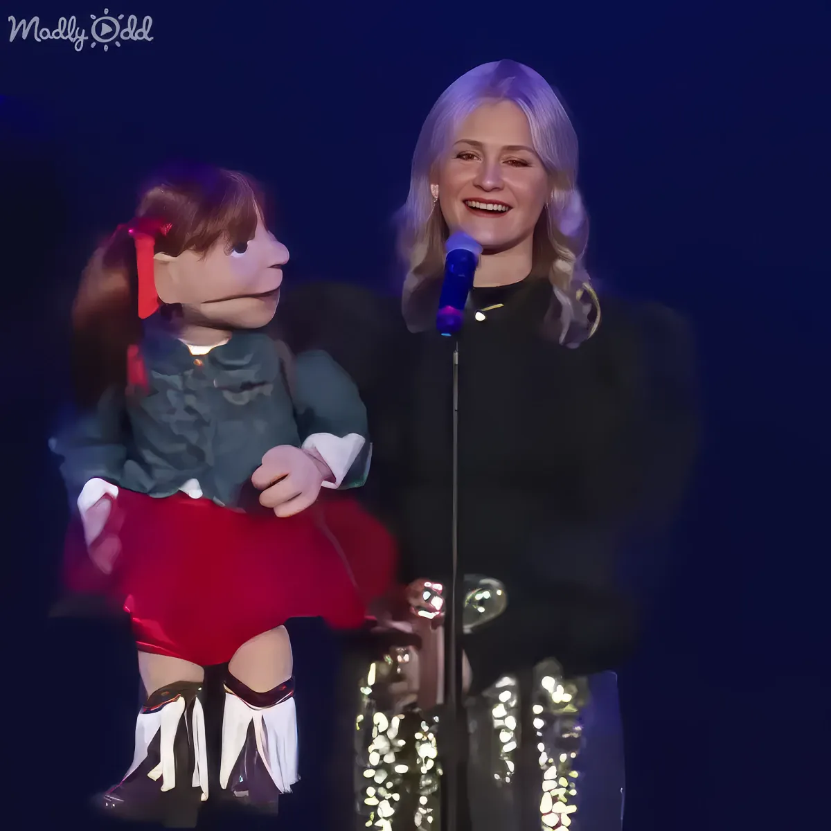 A person stands on stage with a puppet, both smiling, in front of a microphone.