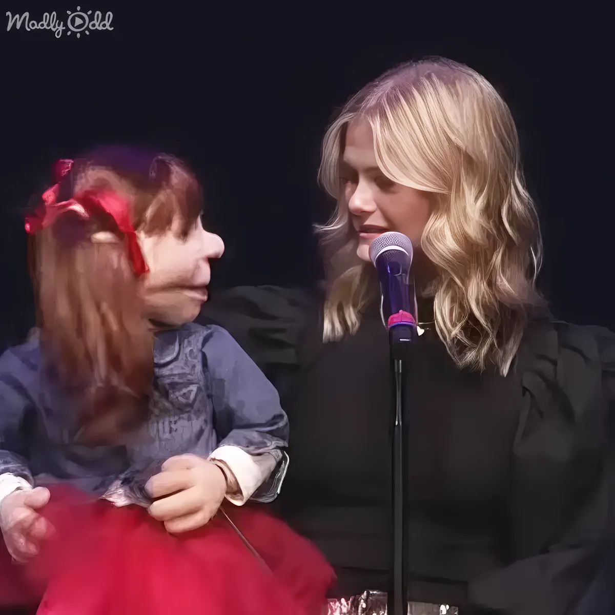 A woman in black speaks into a microphone while a young girl in a red dress holds a ribbon.